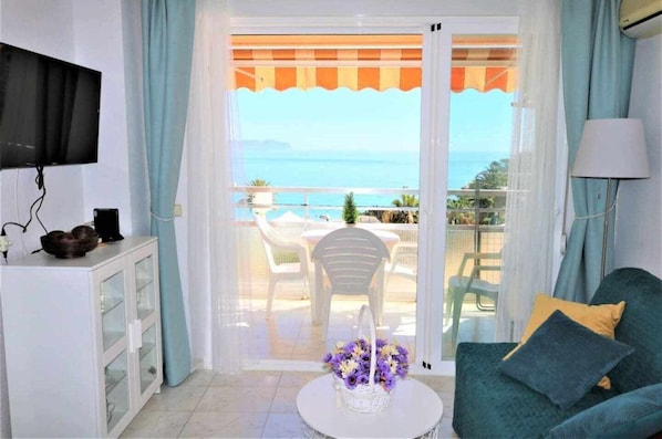 A room with a TV, a white cabinet, a green sofa, a small table with a vase of flowers, and a balcony with a table and chairs overlooking the sea.