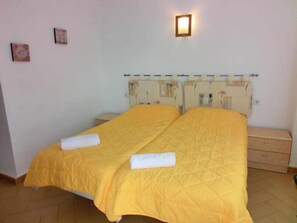 Un lit double avec une couette jaune, deux serviettes blanches, une table de chevet et un luminaire fixé au mur.