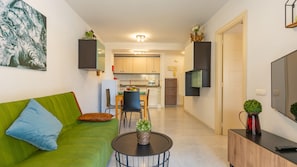 A compact living space with a green sofa, a dining table, a kitchen area, and a TV mounted on the wall.
