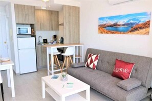 A compact living space with a kitchenette, a sofa with decorative pillows, and a painting of a mountainous landscape.