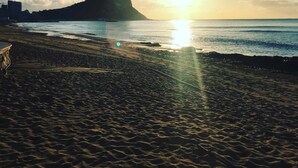 Ein Strand mit einer großen Felsformation im Hintergrund, die Sonne geht über dem Meer unter, und der Himmel ist wolkenlos.