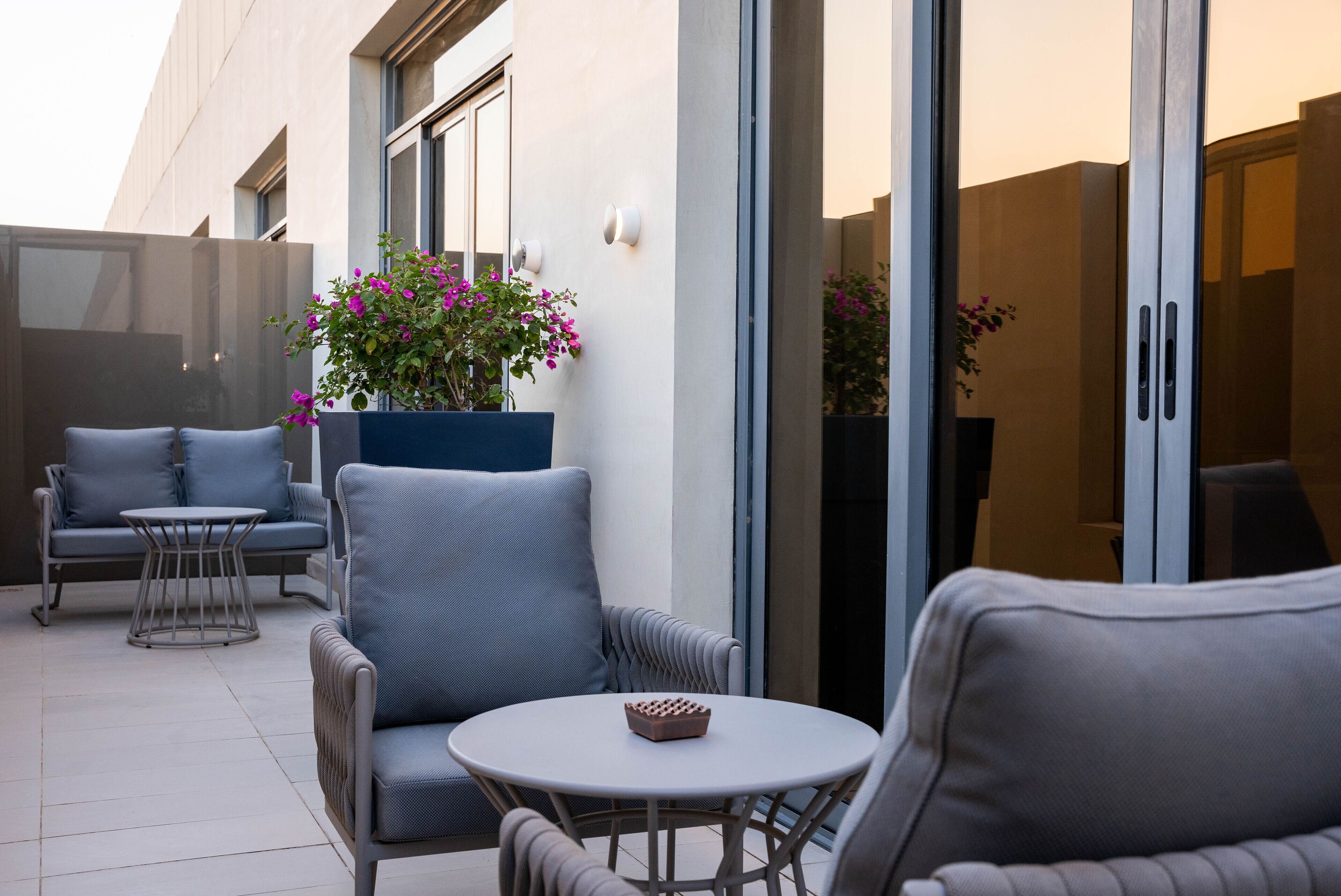 A modern outdoor seating area with grey upholstered chairs and a round table, a planter with pink flowers, and a building with large glass doors.