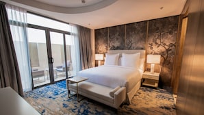 Habitación de hotel con una cama grande, dos mesitas de noche con lámparas, un pequeño sofá y vistas a un área exterior por una puerta corrediza de vidrio.