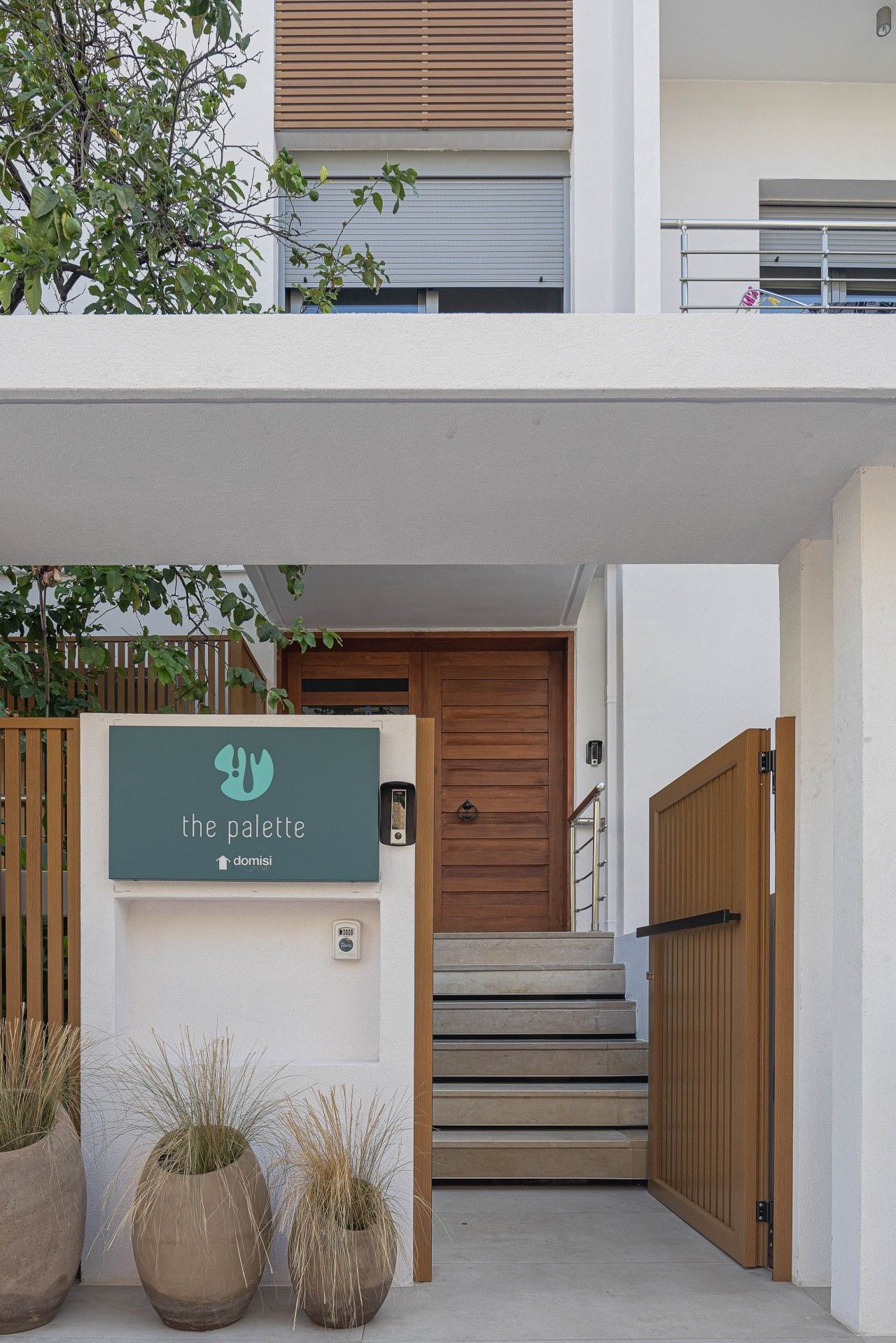 A modern building entrance with a wooden door, a sign for 'the palette,' and potted plants.