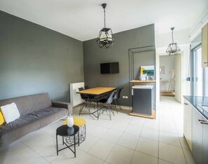 A modern living room with a sofa, a dining table, and chairs. There is a TV mounted on the wall, and a kitchen area is visible in the background.