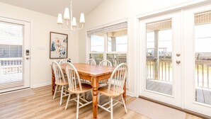 A dining area with a wooden table and white chairs, a chandelier, and large windows with blinds.