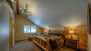 A bedroom with a sloped ceiling, a wooden bed, a nightstand with a lamp, and a window with a view of trees.