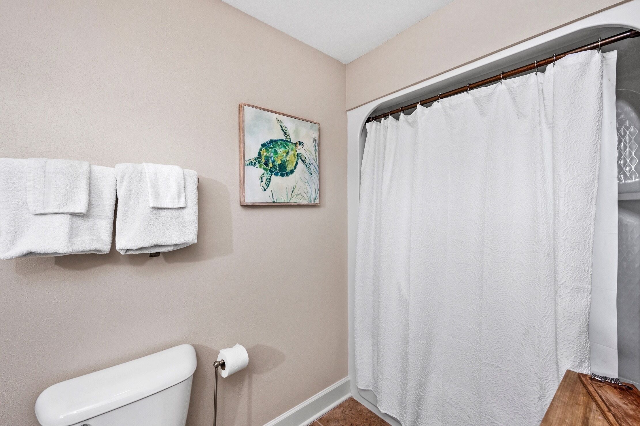 Une salle de bain avec des toilettes, un rouleau de papier toilette, deux serviettes blanches accrochées au mur, une œuvre d’art encadrée représentant une tortue et un rideau de douche blanc.