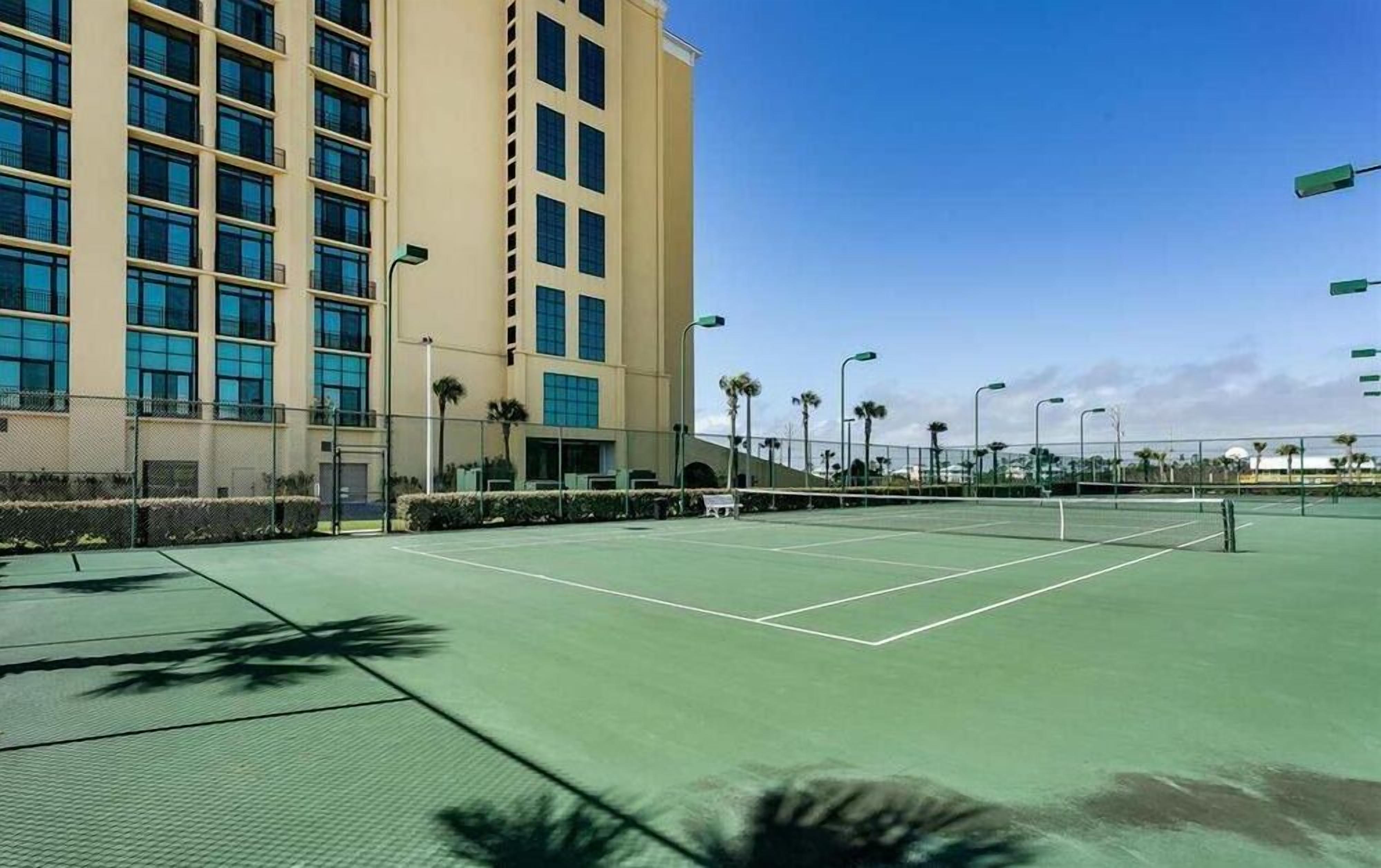 A tennis court with a net, surrounded by a fence and palm trees, adjacent to a multi-story building with large windows.