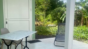 A covered patio with a table and chairs, a door, and a view of a garden with trees and plants.