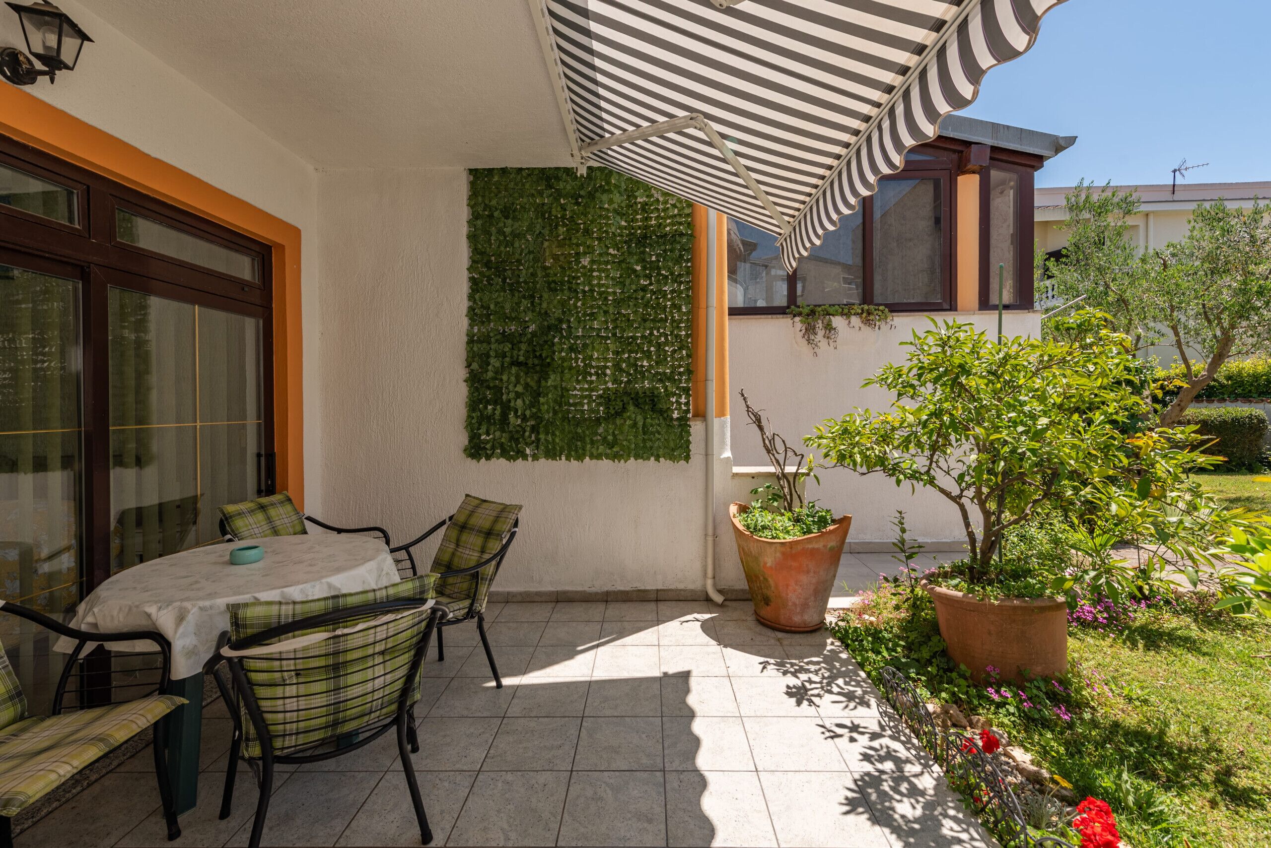 A patio with a table and chairs, a striped awning, a green wall, and potted plants.