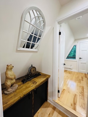 Cozy retreat with a wooden floor, a door leading to another room, a small table with a bear figurine and clock, and a large arched window.