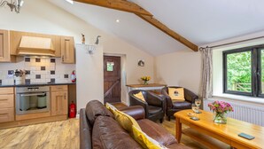 A modern kitchen with wooden cabinets, a built-in oven, and a red fire extinguisher. There is a brown leather sofa with yellow cushions, a wooden dining table with a vase of flowers, and a window with a view of greenery. - Stables Cottage - deep in the Devon countryside (Holsworthy)