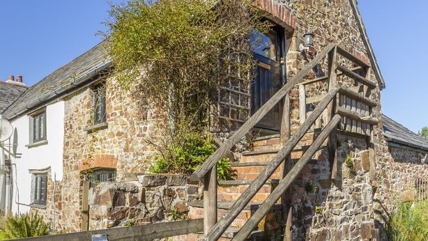 A stone building with a wooden staircase leading to a door, surrounded by a gravel path and greenery. - Appleloft Suite in 17th century Farmhouse (Holsworthy)
