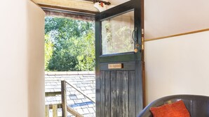 A room with a wooden floor, a dark brown leather sofa with a red cushion, and a door leading to an outdoor area with a wooden deck and a view of trees. - Appleloft Suite in 17th century Farmhouse (Holsworthy)