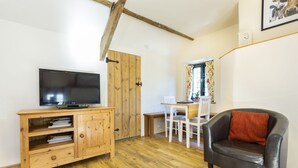A living room with a wooden TV stand, a leather sofa, a dining table with chairs, and a wooden door. - Appleloft Suite in 17th century Farmhouse (Holsworthy)