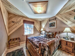 Ein Schlafzimmer mit geneigter Decke, Holzbalken, einem großen Fenster, einem Bett mit gemustertem Überwurf, einem Nachttisch mit Lampe und einer Kommode.