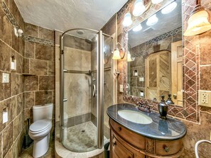 Ein Badezimmer mit einer gläsernen Duschkabine, Toilette, Waschbecken mit Marmorplatte und einem großen, beleuchteten Spiegel.