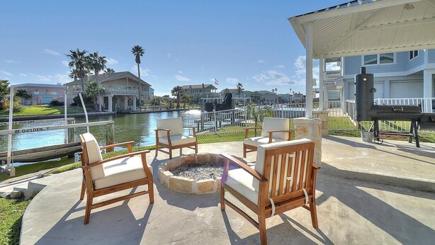 An outdoor patio with wooden chairs and a fire pit, overlooking a canal with buildings and palm trees in the background.