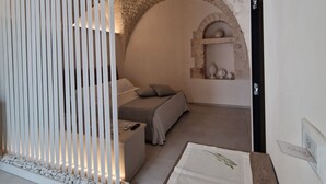 Une chambre moderne et minimaliste, dotée d’une arche en pierre, d’un lit recouvert d’une couette à rayures grises et blanches, d’une cloison blanche à lattes verticales et d’une petite étagère ornée d’objets décoratifs.