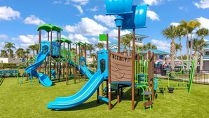 Un parque infantil con toboganes y estructuras para escalar, rodeado de palmeras y un cielo despejado.