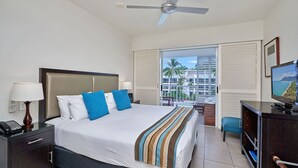 A hotel room with a bed, a desk, a television, a ceiling fan, and a view of a building and palm trees. - Beach Club Top Floor Spa Suite 3131 (Palm Cove)