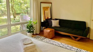 A cozy room with a dark green sofa, a white knitted rug, and a wooden bench. A large window offers a view of greenery outside. - Mid-Century Gem • Creekside • King Beds • Near UNC (Chapel Hill)