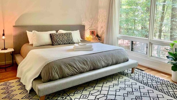 A modern bedroom with a large bed, a bedside table, a nightstand, and a window with a view of trees. - Mid-Century Gem • Creekside • King Beds • Near UNC (Chapel Hill)