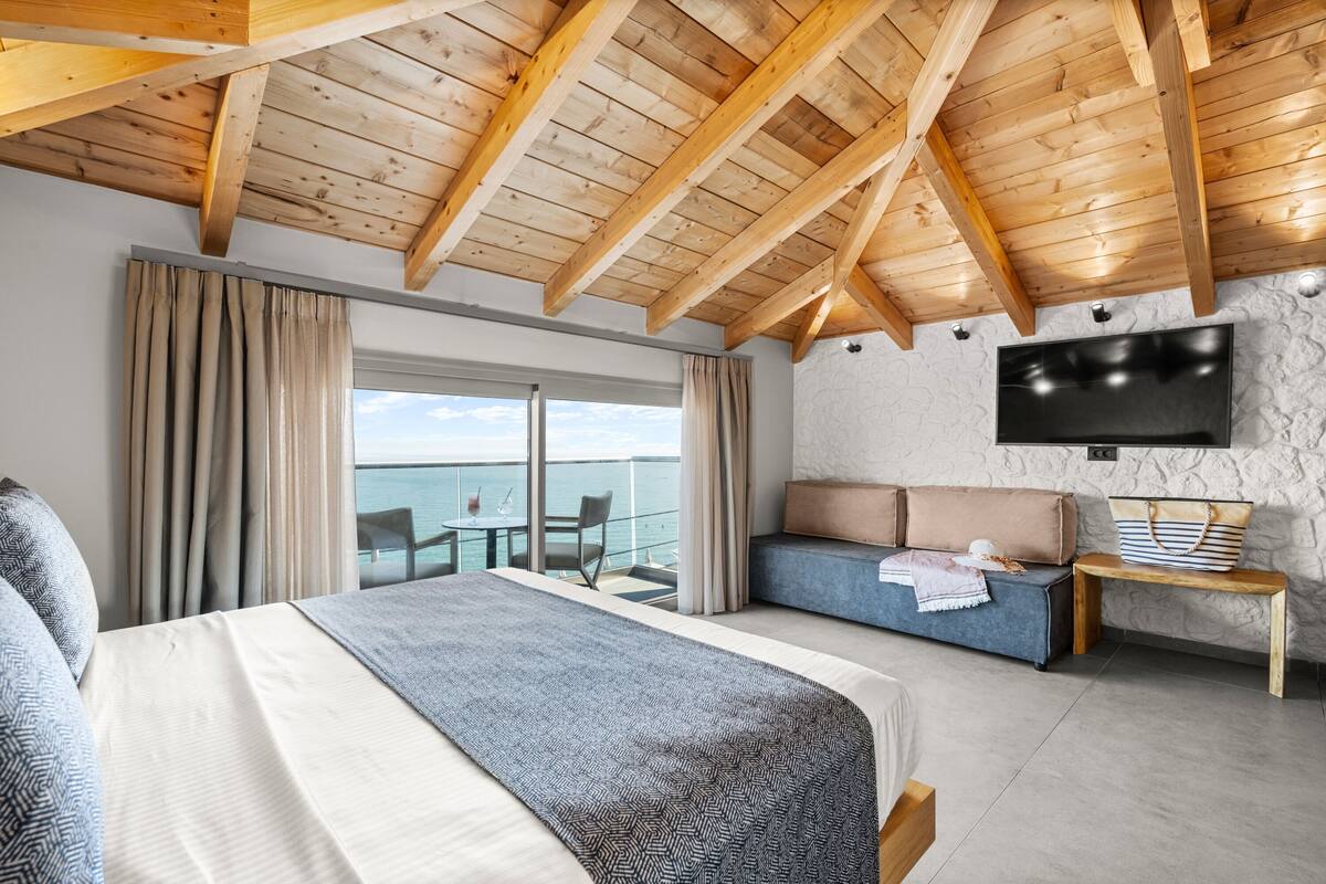 A hotel room with a large bed, a sofa, a TV, a balcony with a view of the sea, and a wooden ceiling.