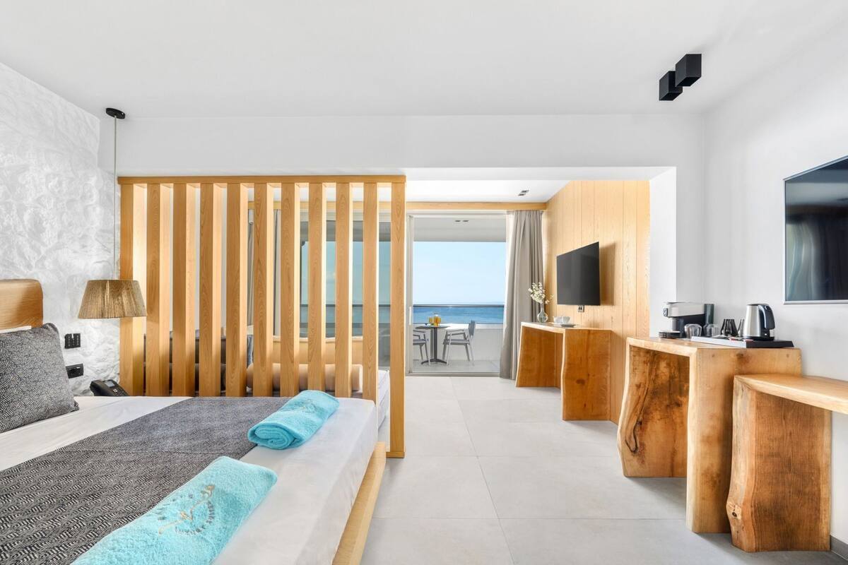 A modern hotel room with a bed, a desk, a TV, and a view of the sea.