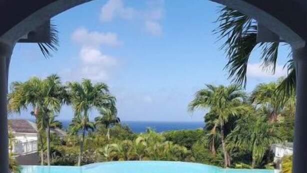 A view through an archway of a swimming pool with a fountain, surrounded by palm trees and a clear sky. - Sugar Hill Resort, St.James (Mount Standfast)