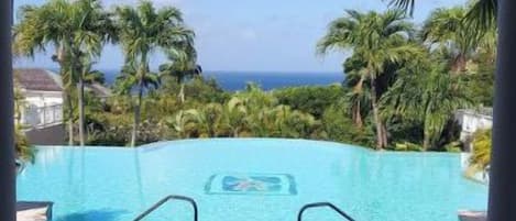 A view through an archway of a swimming pool with a fountain, surrounded by palm trees and a clear sky.