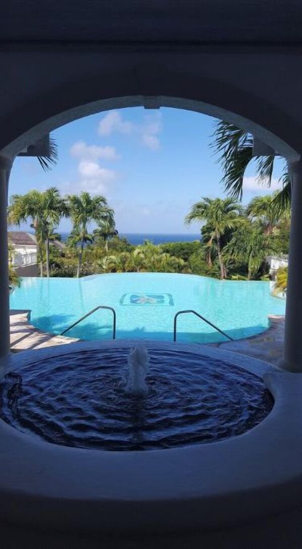 A view through an archway of a swimming pool with a fountain, surrounded by palm trees and a clear sky.