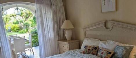 A bedroom with a bed, a nightstand, a ceiling fan, a window with curtains, and a view of a garden.