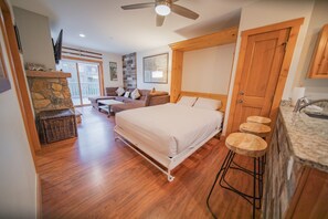 A spacious room with a large bed, a wooden ceiling fan, a stone fireplace, a balcony with a view, and a kitchen area with stools.