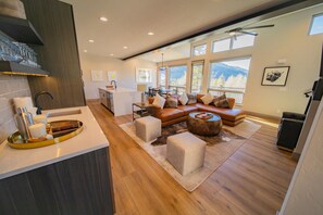 Ein modernes Wohnzimmer mit Sofa, Hocker und Couchtisch. Es gibt eine Küchenzeile mit Spüle und Arbeitsplatte. Der Raum hat große Fenster und Blick ins Freie.