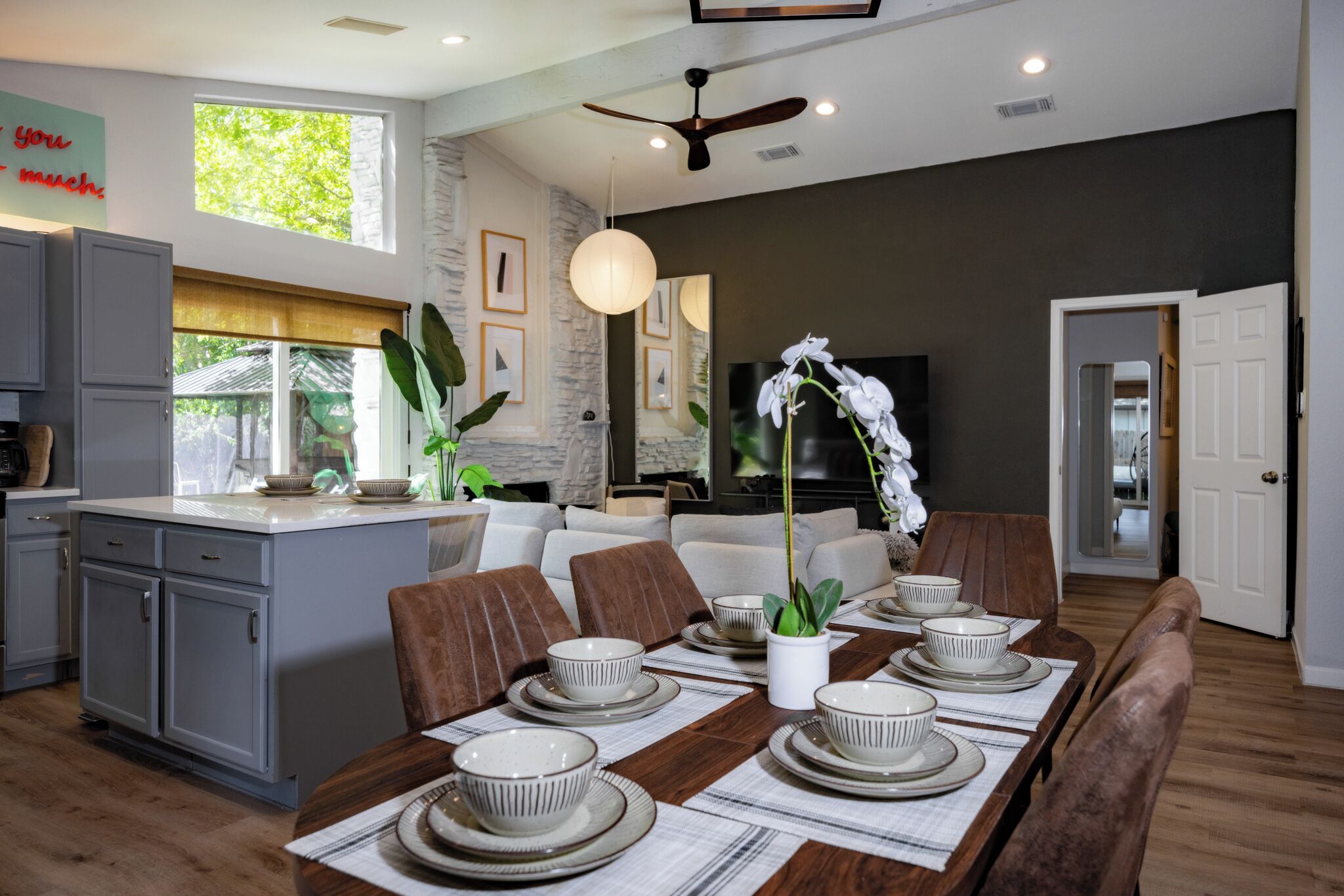 Une cuisine moderne avec des meubles gris, un coin repas avec une table en bois dressée pour six personnes, et un salon avec un canapé et un téléviseur.
