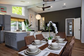A modern kitchen with grey cabinets, a dining area with a wooden table set for six, and a living room with a sofa and a TV.