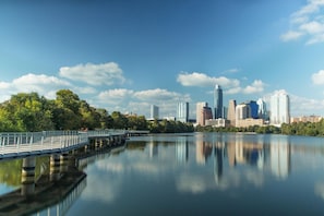 A city skyline with modern high-rise buildings, a calm lake reflecting the structures, and a bridge with a railing on the left.