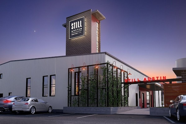 A Still Austin whiskey distillery building with a prominent sign, a parking lot with cars, and a clear sky.