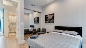 A modern bedroom with a large bed, a chair, a desk, and a bathroom visible through an open door. - Modern 4 Bedroom Charmer w/ Rooftop Deck House #2  (Los Angeles)