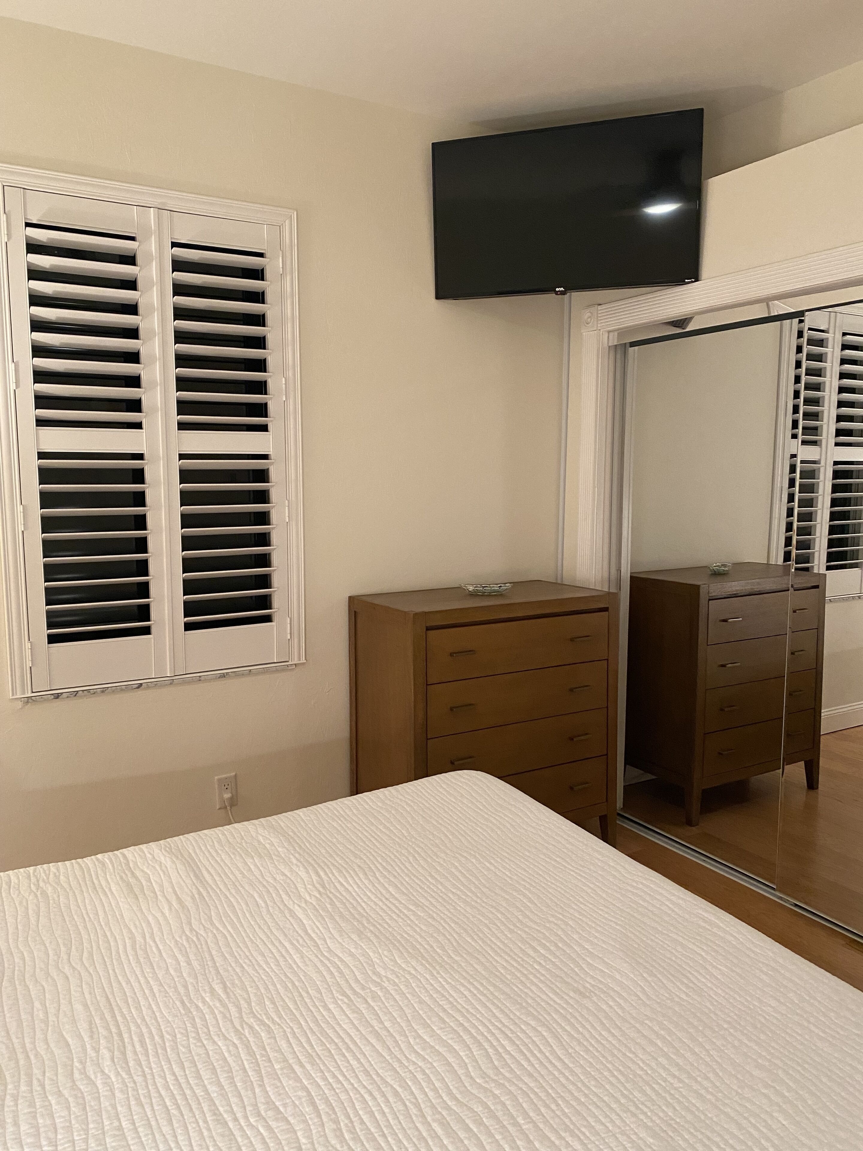Een slaapkamer met een bed, een dressoir, een aan de muur bevestigde televisie en een raam met zonwering.