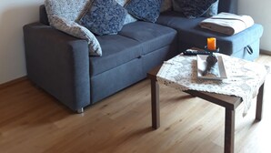 Ein Wohnzimmer mit grauem Sofa, gemusterten Kissen, einem Gemälde, einer Lampe, einem kleinen Tisch mit Kerze und Holzboden.