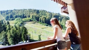 Two people standing by a balcony railing overlooking a forested valley.