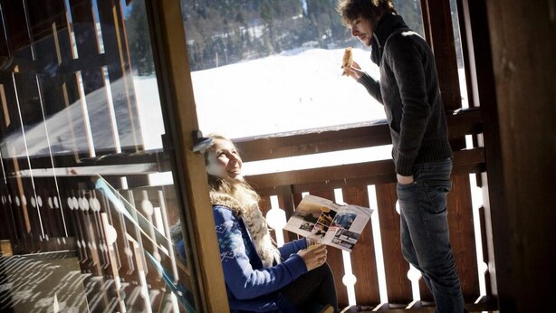 A person standing on a wooden deck, holding a sandwich, and another person sitting indoors reading a magazine.
