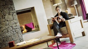 A person sitting on a wooden chair reading a magazine in a room with a stone wall, a fireplace, and a pink rug.