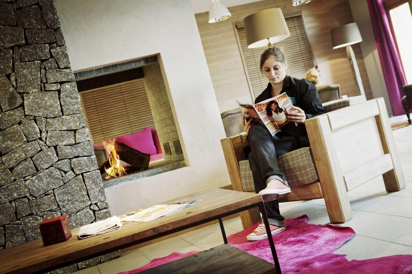 A person sitting on a wooden chair reading a magazine in a room with a stone wall, a fireplace, and a pink rug.