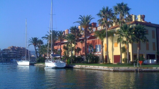 A marina with sailboats docked, palm trees, and a multi-story building with balconies. - Luxury apartment with lovely views over Marina (Marina de Sotogrande, San Roque)