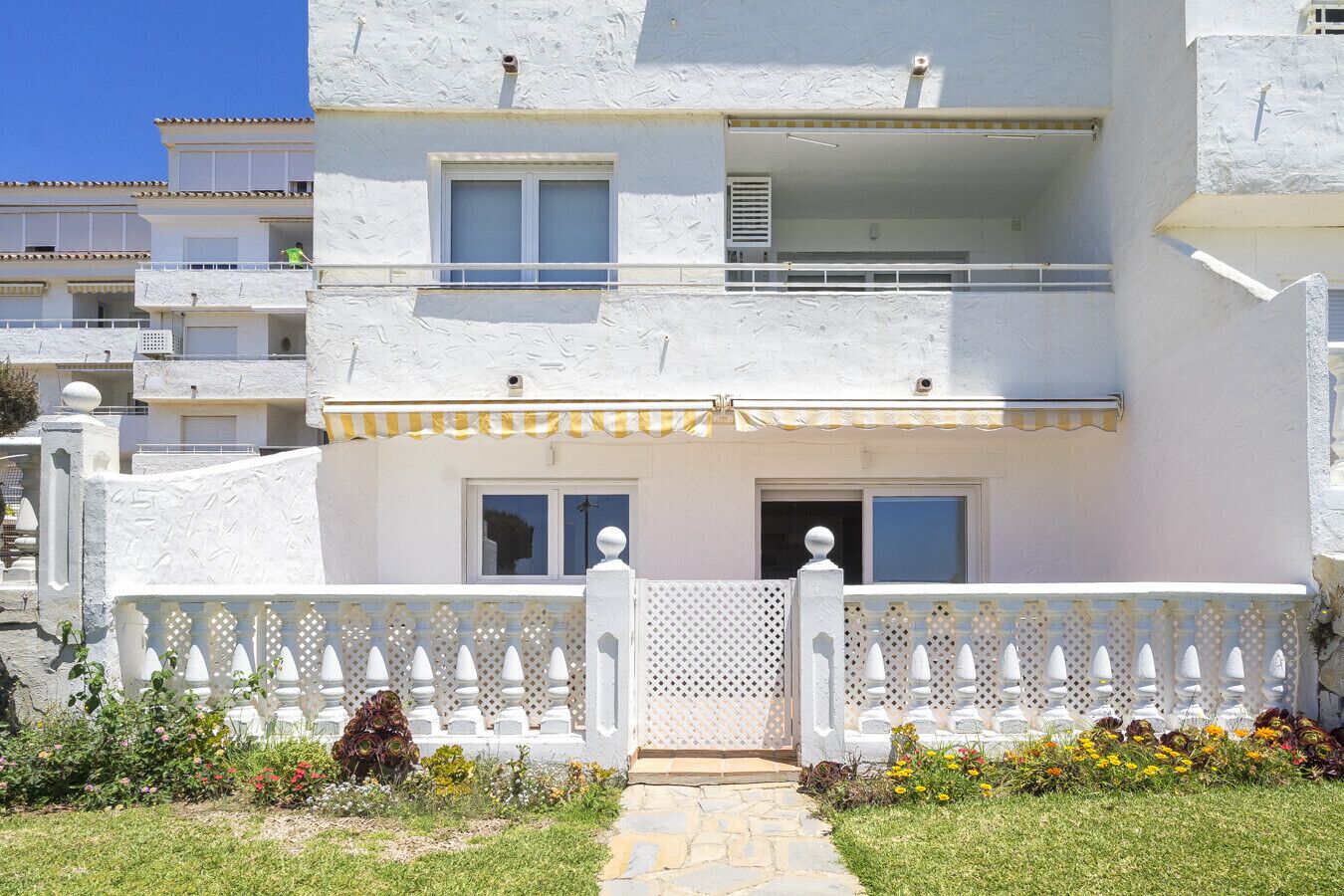 A white building with a balcony, a gated entrance, and a small garden with flowers.