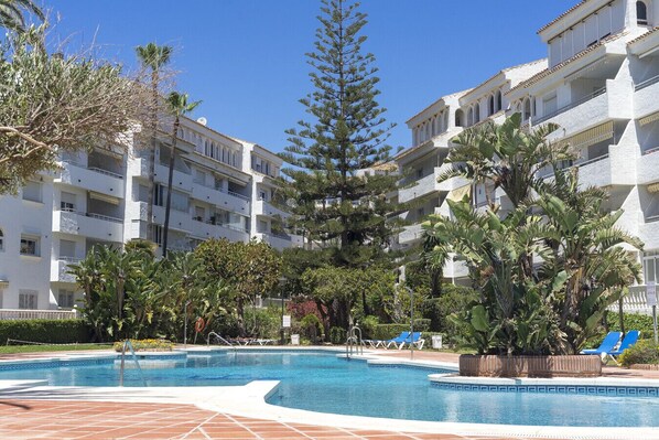 A pool area with a clear sky, surrounded by multi-story buildings with balconies, and various trees and plants.